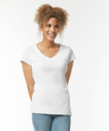 A person with curly hair stands against a plain white background, wearing a white Gildan GD078 V-neck T-shirt and blue jeans, smiling while looking slightly to the side.