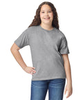 A smiling youth with long curly hair poses against a white background, wearing the Gildan light cotton youth t-shirt in grey and blue jeans, with one hand on their hip.