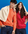 A man in an orange sweatshirt and a woman in a red hoodie, both with dark hair, stand against a blue background. A cream-colored GD101 from Gildan is draped over their heads.