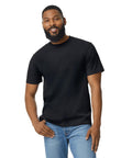 A bearded man stands smiling with one hand on his thigh, wearing a Gildan Softstyle midweight adult no-label enzyme wash black T-shirt and blue jeans against a plain white background.