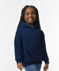 A smiling young girl with braided hair wearing a navy blue hoodie and jeans stands against a plain white background, showcasing the GD57B by Gildan.