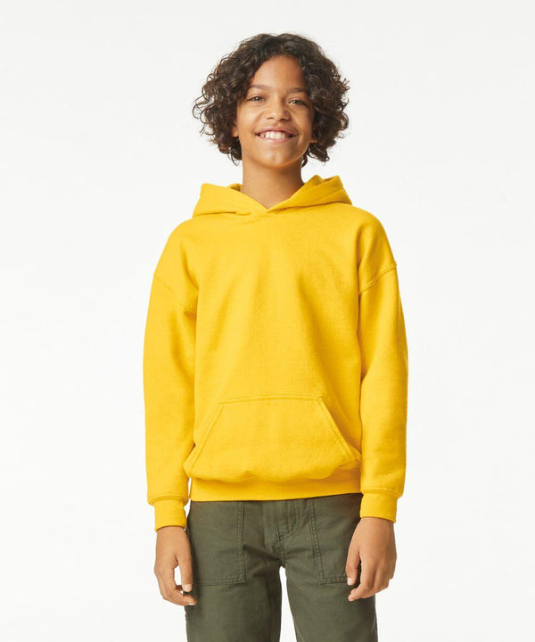 A young person with curly hair stands smiling, wearing a yellow GD67B hoodie by Gildan and green pants.