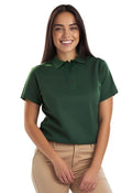 A woman with long dark hair is smiling, wearing an Eco Poloshirt in green and beige pants. They stand against a plain white background, holding their hands in front.