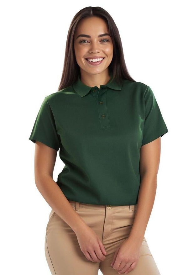 A woman with long dark hair is smiling, wearing an Eco Poloshirt in green and beige pants. They stand against a plain white background, holding their hands in front.