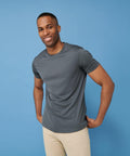 A person stands against a blue background, smiling and wearing a grey Henbury HB024 t-shirt and beige pants.