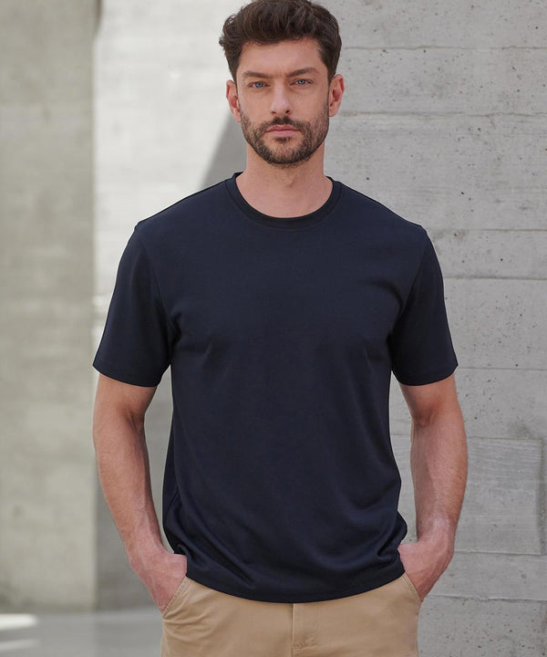 A man with short dark hair and a beard stands indoors against a concrete wall, wearing a Henbury Coolplus wicking t-shirt in black and tan trousers, with his hands in his pockets.