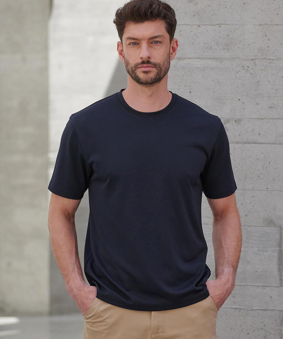 A man with short dark hair and a beard stands indoors against a concrete wall, wearing a Henbury Coolplus wicking t-shirt in black and tan trousers, with his hands in his pockets.