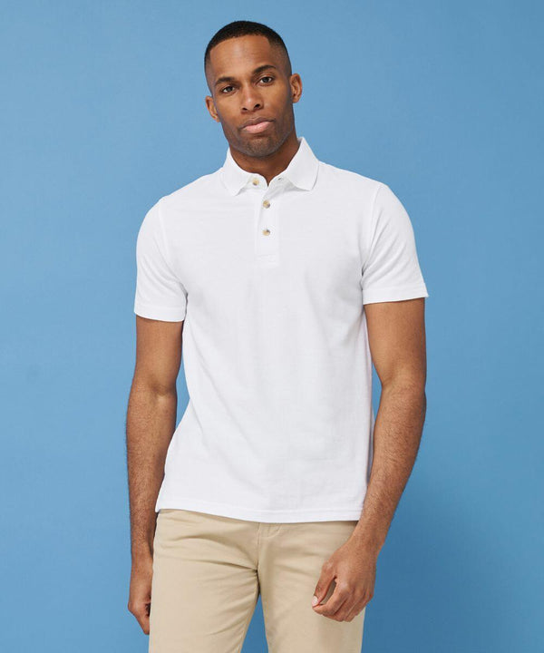 A man wearing a white Henbury HB100 polo shirt and beige pants stands against a blue background.