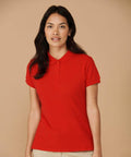Person with long dark hair wearing a red short-sleeve Henbury HB102 polo shirt and beige pants stands against a beige background.