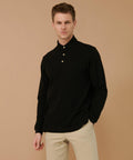 A person stands against a beige background wearing a black long-sleeve Henbury HB105 polo shirt and beige pants, with a neutral expression.