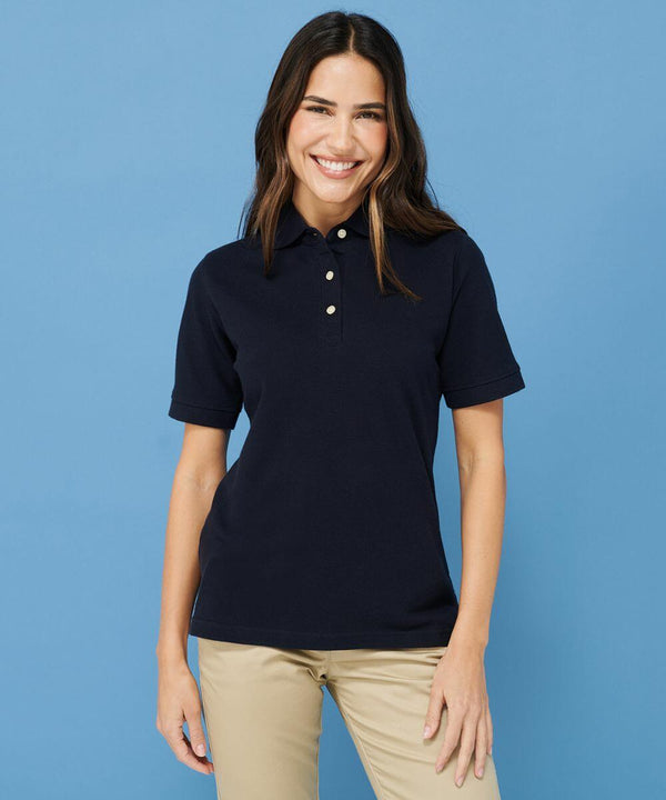 A person with long hair smiling while wearing a navy Henbury HB121 polo shirt and beige pants stands against a blue background.