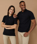Man and woman standing side by side, both wearing Henbury HB305 black polo shirts and beige pants, smiling against a brown background.