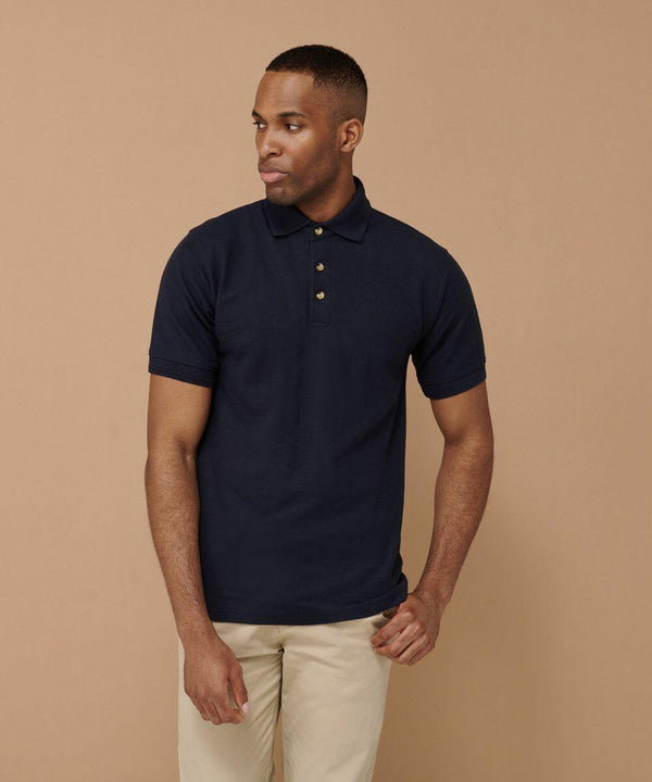 A man stands confidently in front of a beige background, wearing a Henbury HB410 navy blue short-sleeve polo shirt and beige pants.