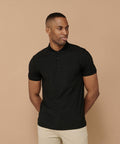 A person wearing a black Henbury HB460 polo shirt and beige pants stands against a beige background with their hands in their pockets, looking to the side.