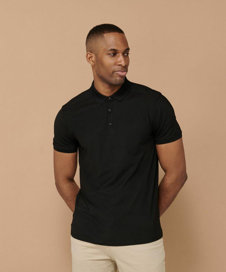 A person wearing a black Henbury HB460 polo shirt and beige pants stands against a beige background with their hands in their pockets, looking to the side.