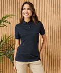 A woman in a Henbury HB466 navy polo shirt and beige pants stands smiling in front of a wooden slat wall with a plant beside her.