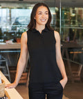 A woman with long dark hair, smiling indoors, wears a Henbury Women's sleeveless Coolplus polo shirt and black trousers. She stands with one hand on a counter and the other in her pocket, with tables and glass walls behind her.