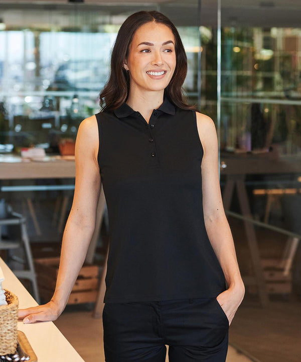 A woman with long dark hair, smiling indoors, wears a Henbury Women's sleeveless Coolplus polo shirt and black trousers. She stands with one hand on a counter and the other in her pocket, with tables and glass walls behind her.