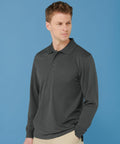 A man wearing a long-sleeved, charcoal-colored Henbury HB478 polo shirt and beige pants stands against a solid blue background. He is looking to the side with a neutral expression.