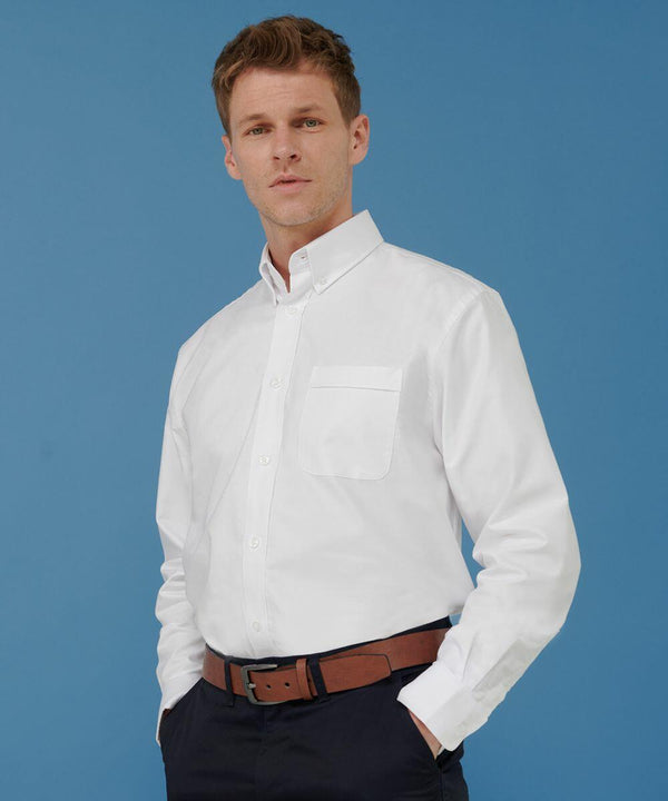 A man wearing a white Henbury HB510 button-up shirt and brown belt stands against a blue background.