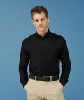 A person stands against a blue background, wearing a Henbury HB512 black button-up shirt, beige pants, and a black belt, with hands in pockets.