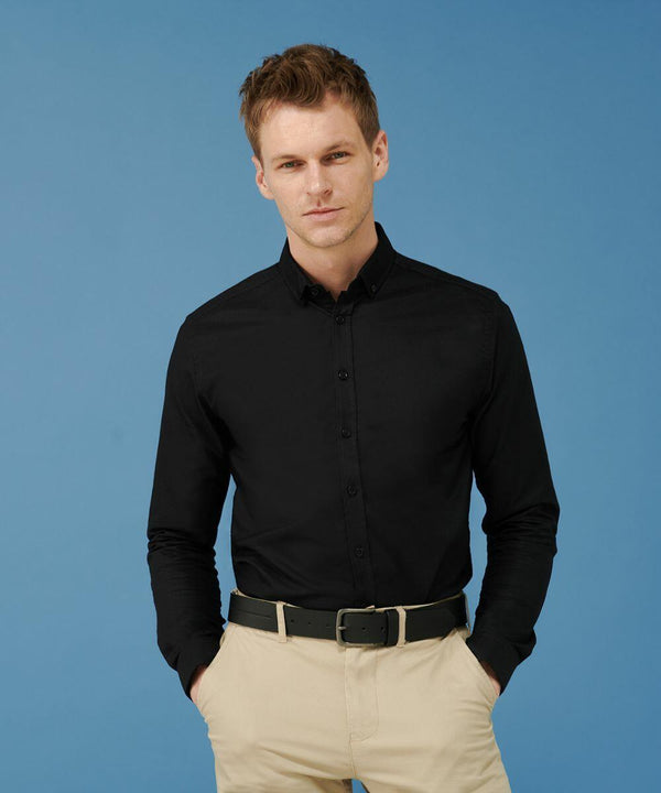 A person stands against a blue background, wearing a Henbury HB512 black button-up shirt, beige pants, and a black belt, with hands in pockets.