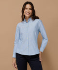 A person is standing against a brown background, wearing a light blue Henbury HB513 button-up shirt and dark pants, and is smiling at the camera.