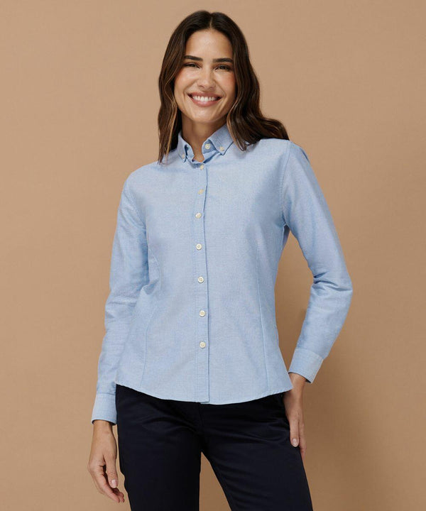 A person is standing against a brown background, wearing a light blue Henbury HB513 button-up shirt and dark pants, and is smiling at the camera.