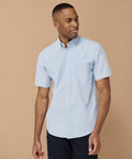 A man stands against a beige background, wearing a light blue Henbury HB515 short-sleeve button-up shirt with a chest pocket and dark pants.