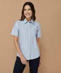 A person with long brown hair wearing a light blue Henbury HB516 short-sleeve button-up shirt and dark pants stands against a beige background, smiling and looking to the right.