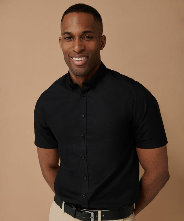A man in a short-sleeved, black Henbury HB517 button-up shirt and beige pants stands against a plain brown background, smiling.