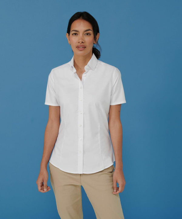 A person wearing a white Henbury HB518 short-sleeve button-up shirt and beige pants stands against a blue background.