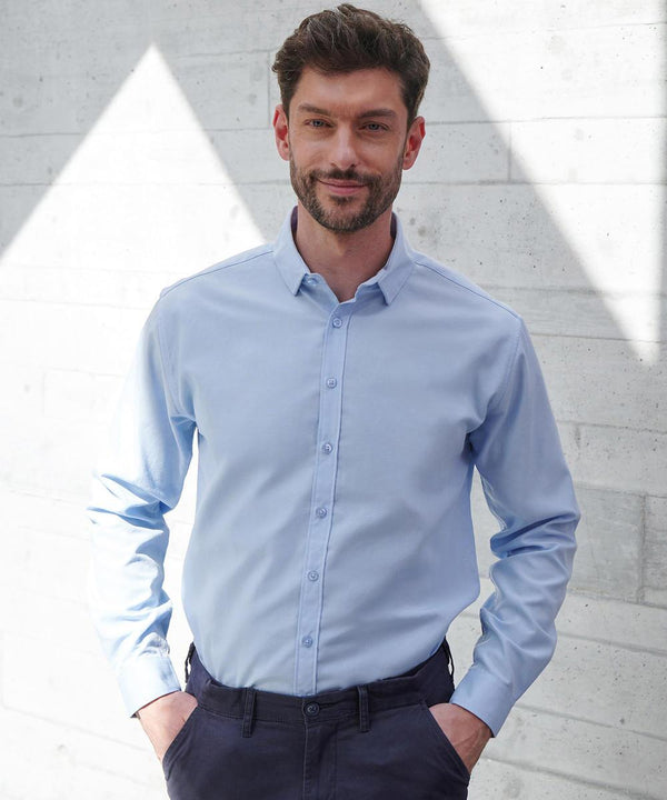 A man with short dark hair and a beard stands against a light concrete wall, wearing a Henbury Long Sleeve Cotton-Feel Coolplus Shirt in light blue and dark trousers, his hands in his pockets.