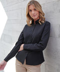 A woman with shoulder-length blonde hair wears a Henbury Women's long sleeve cotton-feel Coolplus shirt in black and beige trousers, standing before a light grey concrete wall with one arm bent and the other extended.