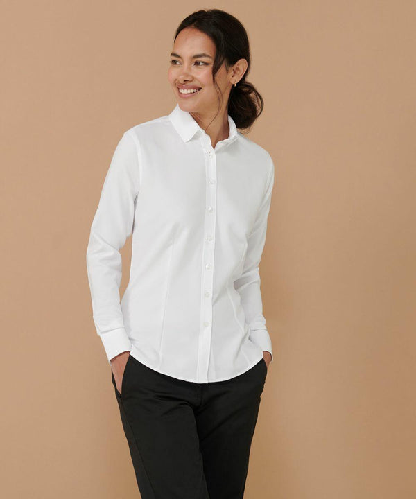 A person is standing with hands in pockets, wearing a button-up white shirt and black pants (HB591 by Henbury), smiling against a plain beige background.