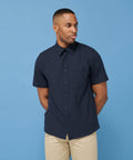 A person wearing a short-sleeve, navy Henbury HB595 and beige pants stands against a blue background, looking off to the side.