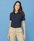 A woman wearing a navy Henbury HB596 short-sleeve button-up shirt and beige pants stands against a blue background, smiling.