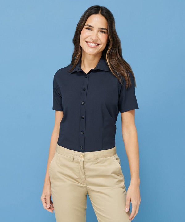 A woman wearing a navy Henbury HB596 short-sleeve button-up shirt and beige pants stands against a blue background, smiling.