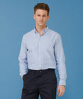 A person stands against a blue background, wearing a light blue Henbury HB640 button-down shirt and dark blue pants.
