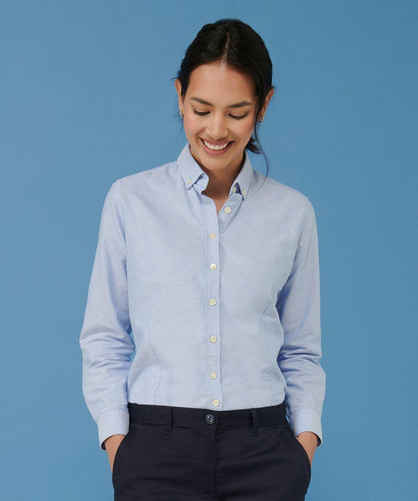 A woman wearing a light blue Henbury HB641 button-up shirt and navy pants looks down and smiles against a blue background.
