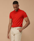 A man wearing a red Henbury HB650 and beige pants stands against a beige background, looking down with one hand in his pocket.