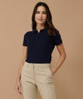 A person with long dark hair is standing against a beige background wearing a Henbury HB651 and beige pants, with one hand in their pocket.