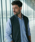 A man with short dark hair and a beard is wearing a Henbury sleeveless zip-through jumper over a light blue shirt. He stands indoors by large windows, looking to the side with a neutral expression.