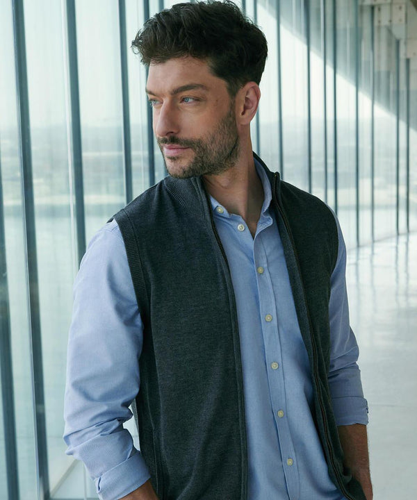 A man with short dark hair and a beard is wearing a Henbury sleeveless zip-through jumper over a light blue shirt. He stands indoors by large windows, looking to the side with a neutral expression.