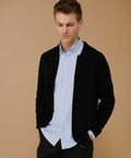 A man wearing a light blue Henbury HB718 button-up shirt and a black jacket stands with his hands in his pockets against a beige background.