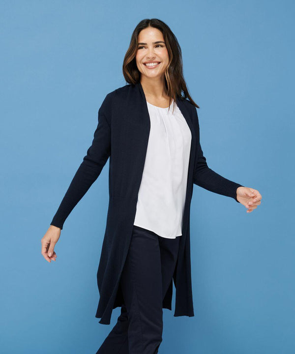 A woman with long dark hair smiles while wearing a white blouse, dark pants, and a long, dark Henbury HB719 cardigan against a blue background.