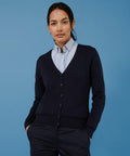 A person wearing a light blue Henbury HB726 button-down shirt underneath a dark blue cardigan stands against a blue background.