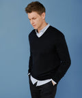 Man with short brown hair wearing a black Henbury HB760 V-neck sweater over a white shirt and black pants stands against a blue background with hands in his pockets, looking down to his left.