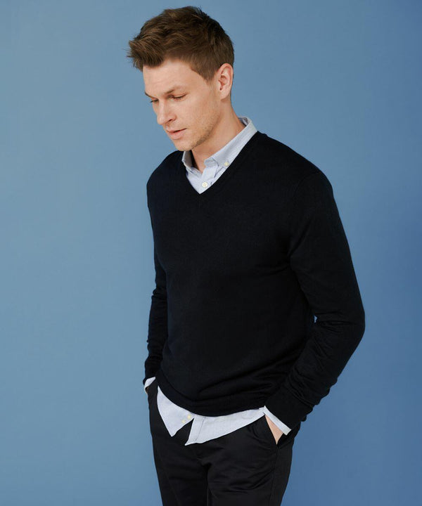 Man with short brown hair wearing a black Henbury HB760 V-neck sweater over a white shirt and black pants stands against a blue background with hands in his pockets, looking down to his left.