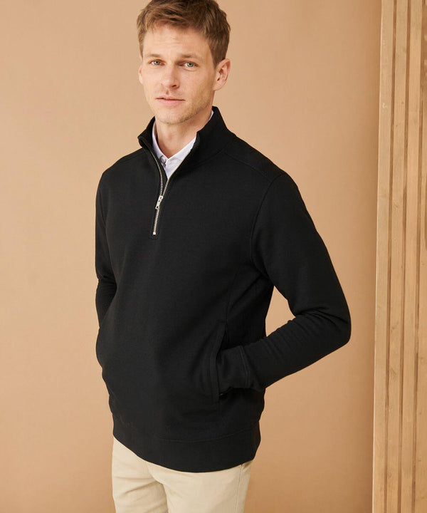 A man stands against a plain background, wearing a black Henbury HB842 and beige pants, with his hands in the pockets of his pullover.